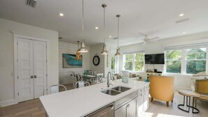 A modern kitchen with a central island, pendant lighting, and a dining area with a sofa and chairs. - Pineapple Paradise~WATER SLIDE/Heated Pool & Spa! (Holmes Beach)