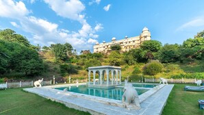 Una piscina con un pabellón y estatuas, rodeada de césped y una colina con un edificio grande al fondo.