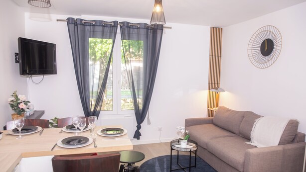 A modern living room with a sofa, a dining table set for four, a TV mounted on the wall, and a window with curtains.