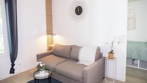 A modern living room with a beige sofa, a round side table with a lamp, a wall clock, and a small potted plant.
