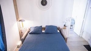 A bedroom with a bed, bedside lamps, a wall clock, and a door leading to another room.