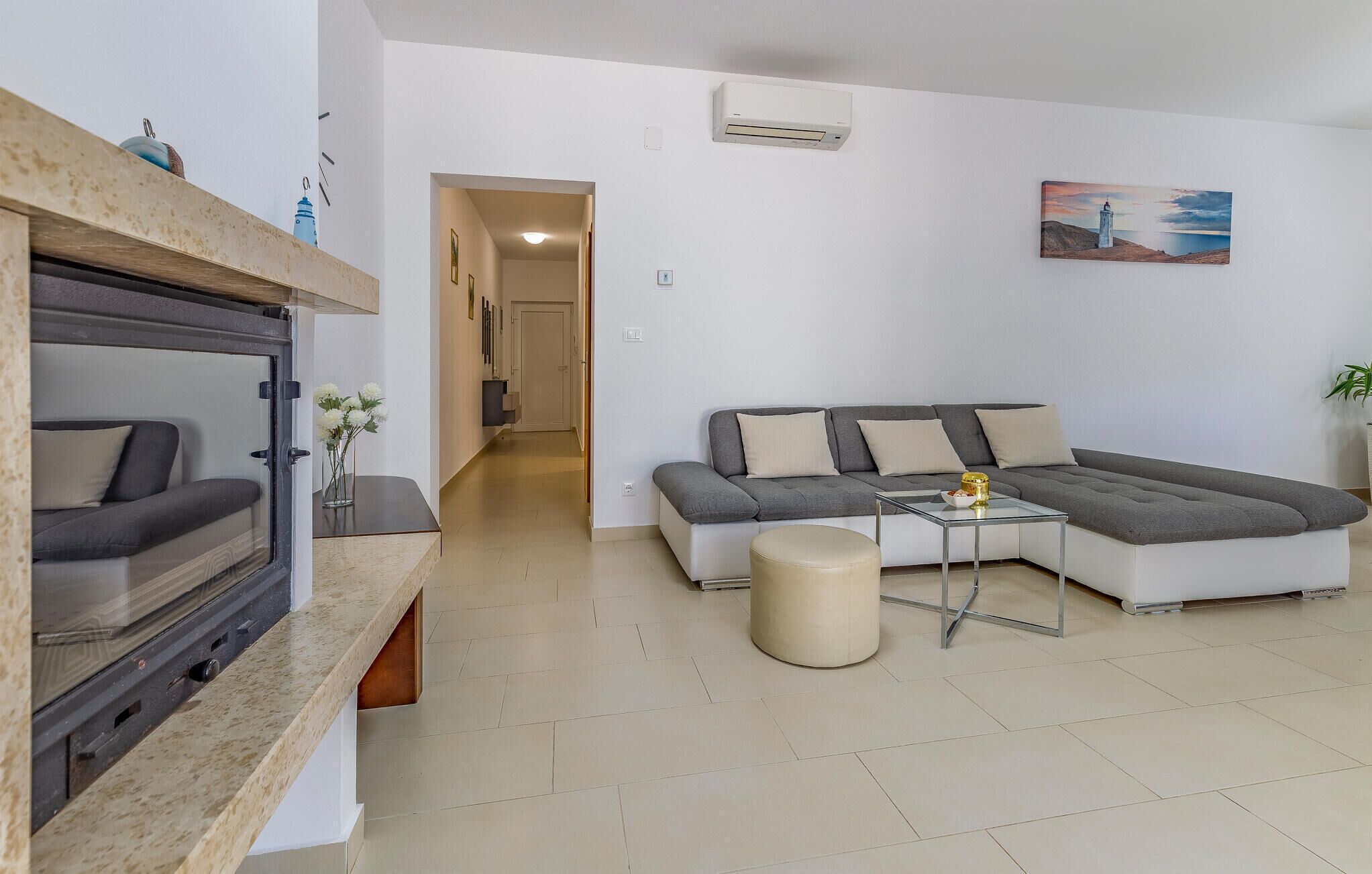 A modern living room with a sectional sofa, a glass coffee table, and a small round ottoman. There is a white air conditioning unit on the wall and a painting above the sofa.