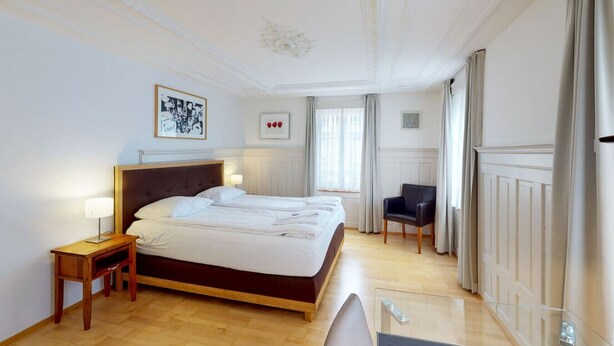 Een hotelkamer met een groot bed, twee nachtkastjes, een stoel en een raam met gordijnen.