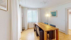 Een eethoek met een houten tafel en stoelen, een raam met witte gordijnen, en een schilderij aan de muur.