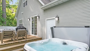 Un bain à remous sur une terrasse en bois, avec deux chaises en bois et un barbecue, à côté d’une maison dotée d’une porte et de fenêtres blanches.