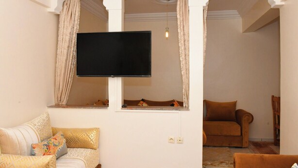 A living room with a flat-screen TV mounted on the wall, two sofas with patterned cushions, and a wooden chair. - Your cozy home in Agadir - discover comfort and tradition (Agadir)