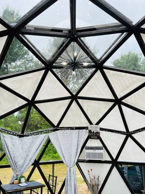 A geodesic dome structure with a central light fixture, a sign reading 'Welcome to our Neck of the Woods,' and a small table with a chair outside.