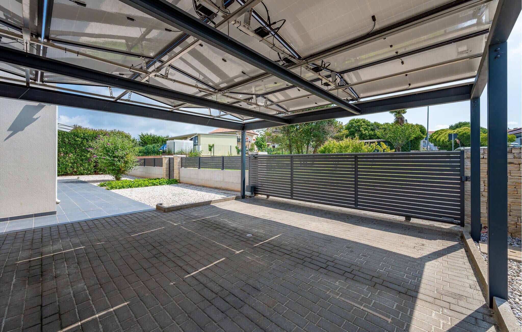 A covered parking area with a tiled floor, metal railings, and a view of greenery and buildings in the background.