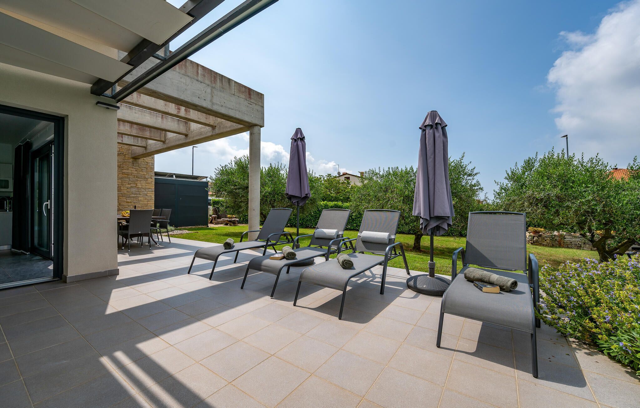 An outdoor patio with six lounge chairs and two umbrellas, a stone wall, and a view of greenery and buildings.