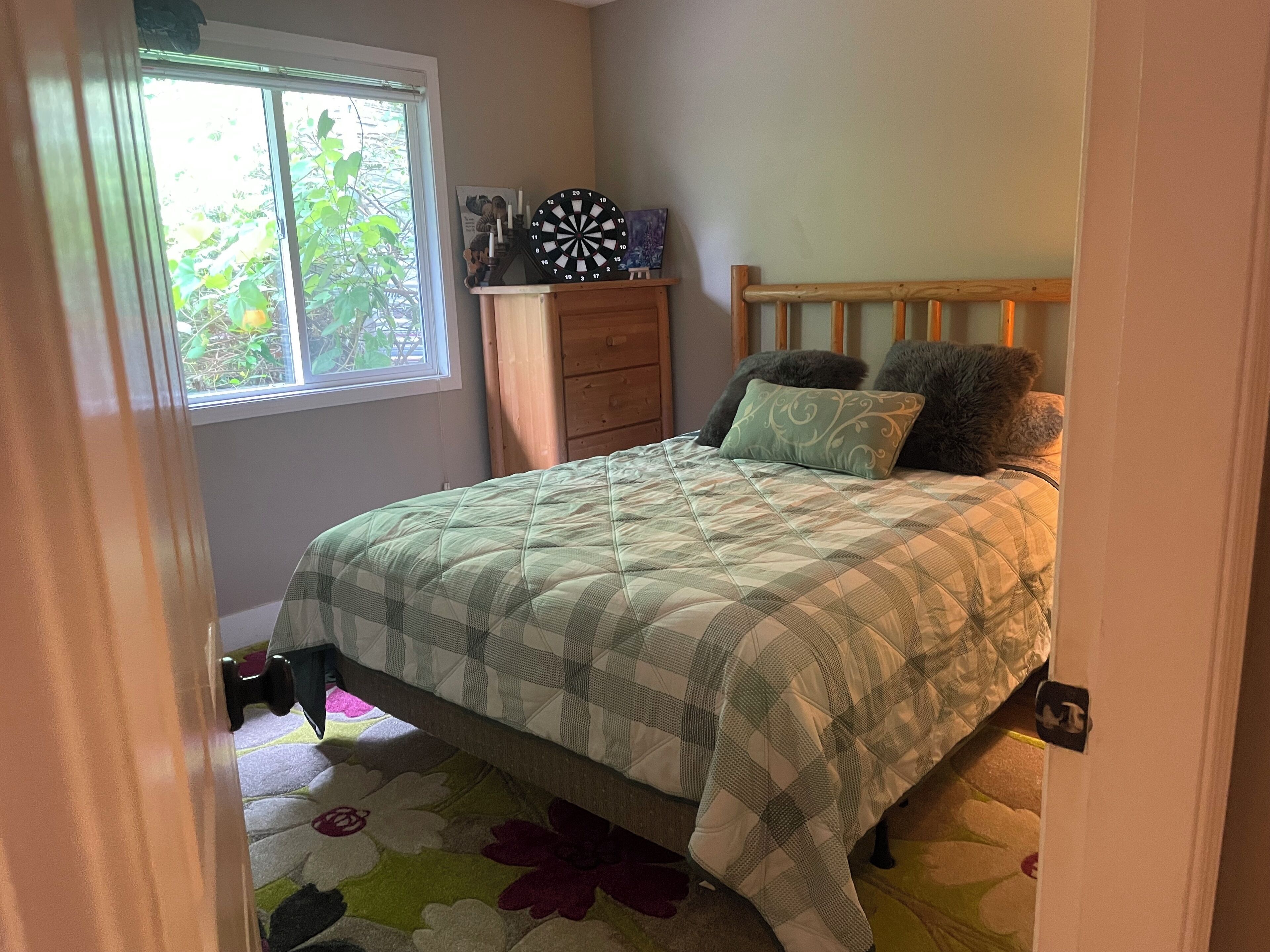 A bedroom with a bed, a dresser, a window, and a dartboard.