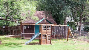 Een houten speelhuisje met een glijbaan, omgeven door een omheind gazon en bomen.