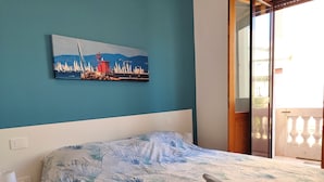 A bedroom with a bed, a nightstand, a lamp, a painting of sailboats, and a balcony. - Colori di Trieste - TriesteCenter (Trieste)
