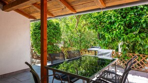 An outdoor patio with a glass-top table and metal chairs, surrounded by lush greenery and a wooden pergola structure.