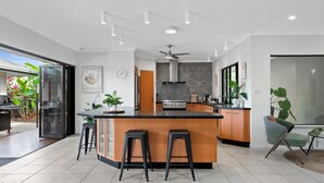 A modern kitchen with a central island, bar stools, a ceiling fan, and a view of the outdoors. - The Pavilion at Trinity Beach (Trinity Beach)