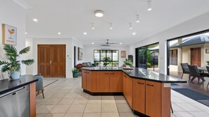 A modern kitchen with wooden cabinets, a black countertop, and a built-in island. There is a dining area with chairs and a table visible through a glass door. - The Pavilion at Trinity Beach (Trinity Beach)