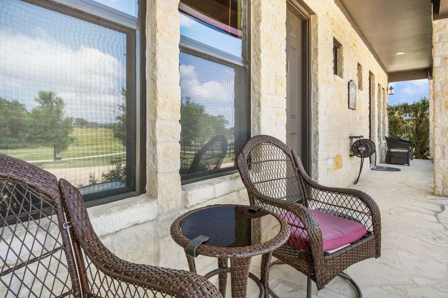 A covered outdoor seating area with wicker chairs and a small table, large windows, and a stone building.