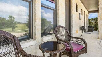 A covered outdoor seating area with wicker chairs and a small table, large windows, and a stone building.