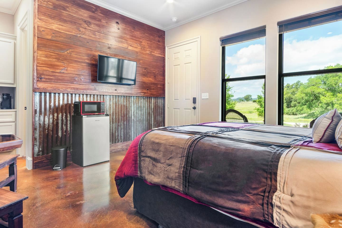 A bedroom with a large bed, a television mounted on a wooden wall, and a view of the outdoors through a large window.