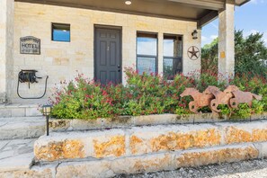 A stone building with a dark door, a sign indicating a hotel, and a decorative metal horse sculpture. - Timbuctoo 2 | Cozy Ranch Suite Near Cowboy Capital (Bandera)