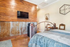 Quarto com parede de madeira, cama com colcha estampada, televisão montada acima da cama e um objeto decorativo na parede.