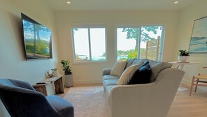 Een moderne woonkamer met een grijze bank, een donkerblauwe fauteuil en een wit vloerkleed. Er zijn twee ramen met uitzicht op het omliggende landschap.