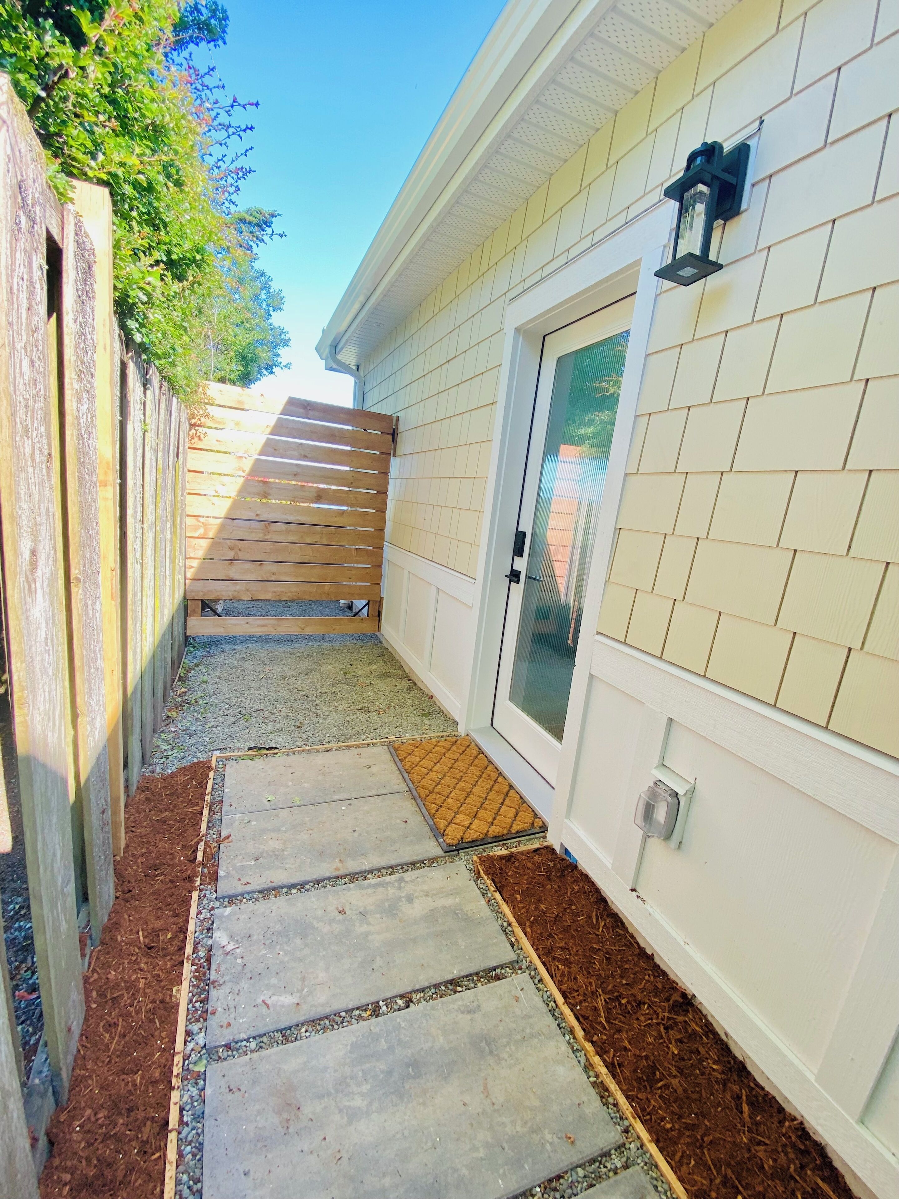 Un étroit sentier mène à une maison. D’un côté, elle est bordée d’une clôture en bois, et de l’autre, elle présente un mur extérieur blanc percé d’une porte et d’une fenêtre.