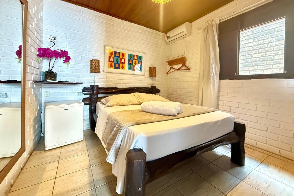 A bedroom with a bed, a desk, a chair, a small refrigerator, and a window with curtains.