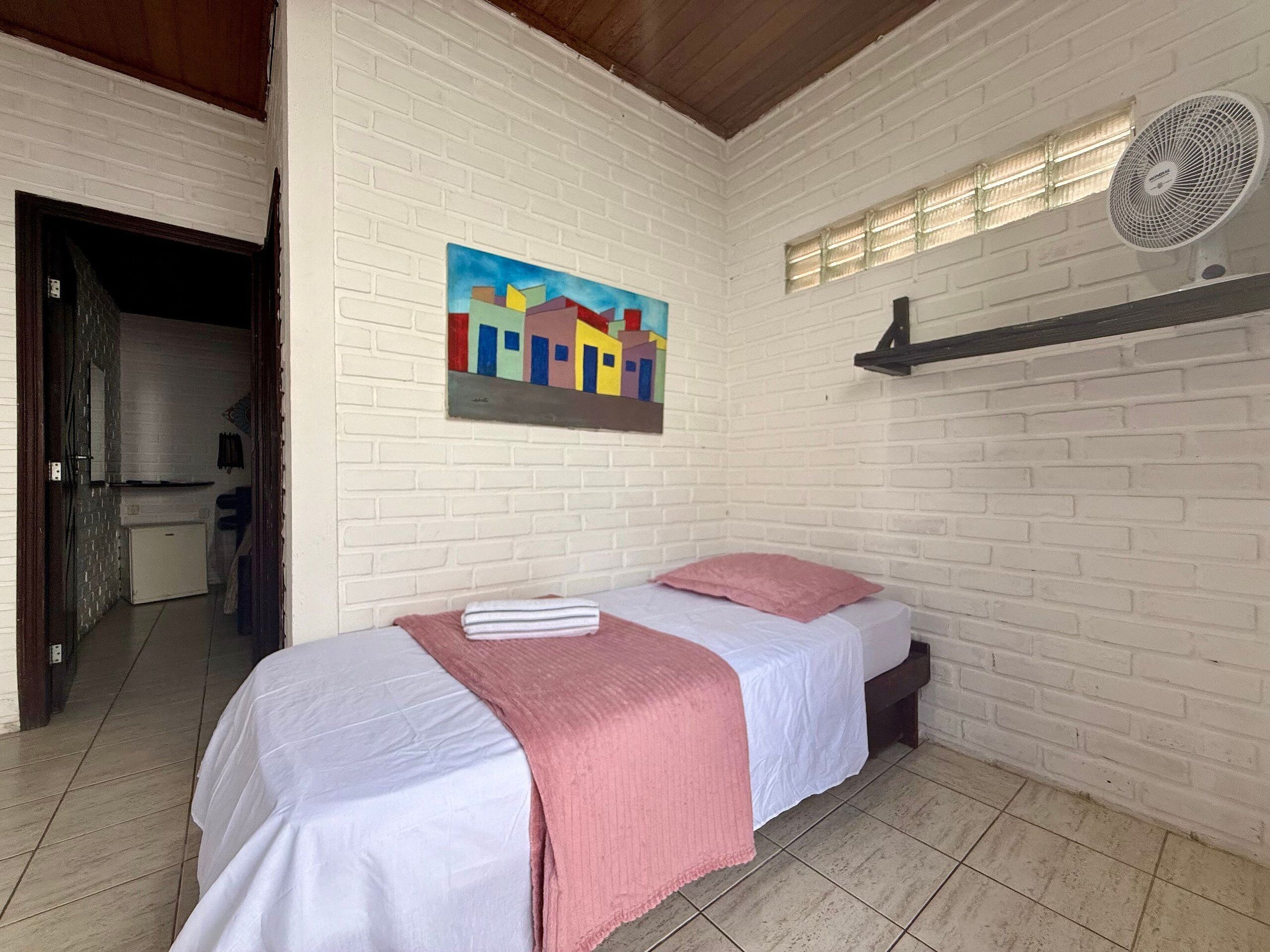 A room with a bed, a white brick wall, a colorful painting, and a ceiling fan.