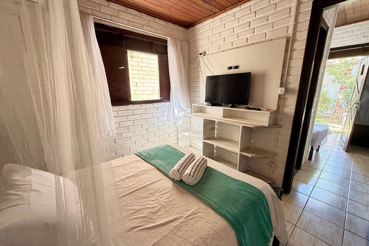 A bedroom with a bed, a TV mounted on a brick wall, a window with curtains, and a door leading to an outdoor area.