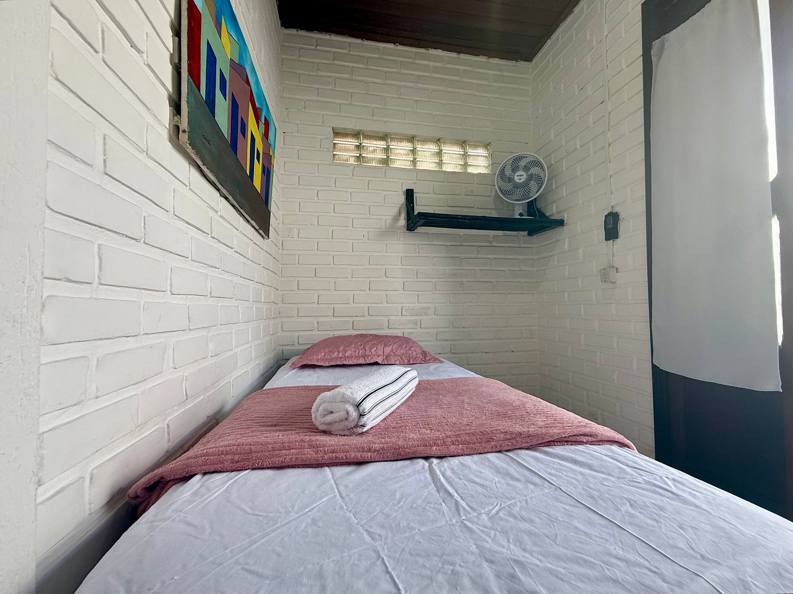 Una habitación pequeña con paredes de ladrillo blanco, una cama con ropa de cama rosa y blanca, una ventana pequeña con persianas y un ventilador de pared.
