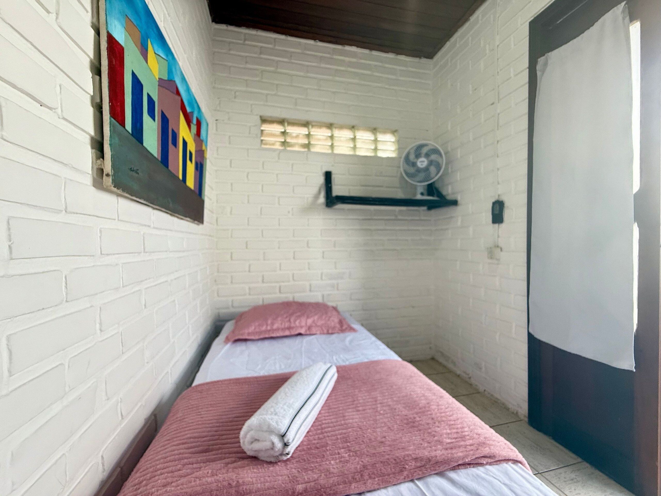 A small, white-walled room with a single bed, a fan, and a colorful painting on the wall.