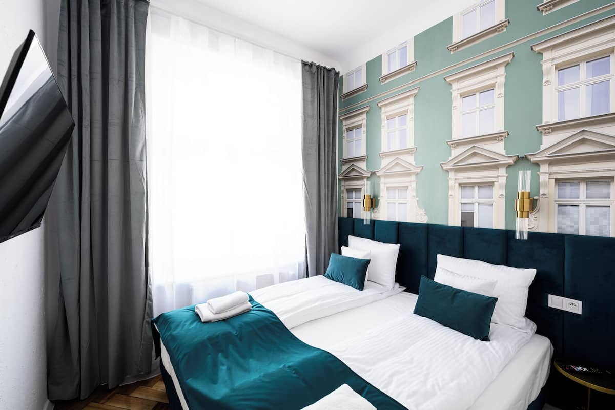 A hotel room with a large bed, grey curtains, a teal bedspread, and a classic architectural wall design.