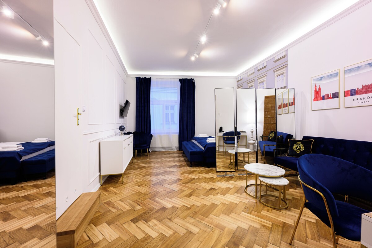 A modern hotel room with a herringbone wooden floor, a blue sofa, a small round table, and a mirror.