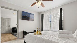 A bedroom with a large bed, a ceiling fan, a painting on the wall, and a window with curtains.