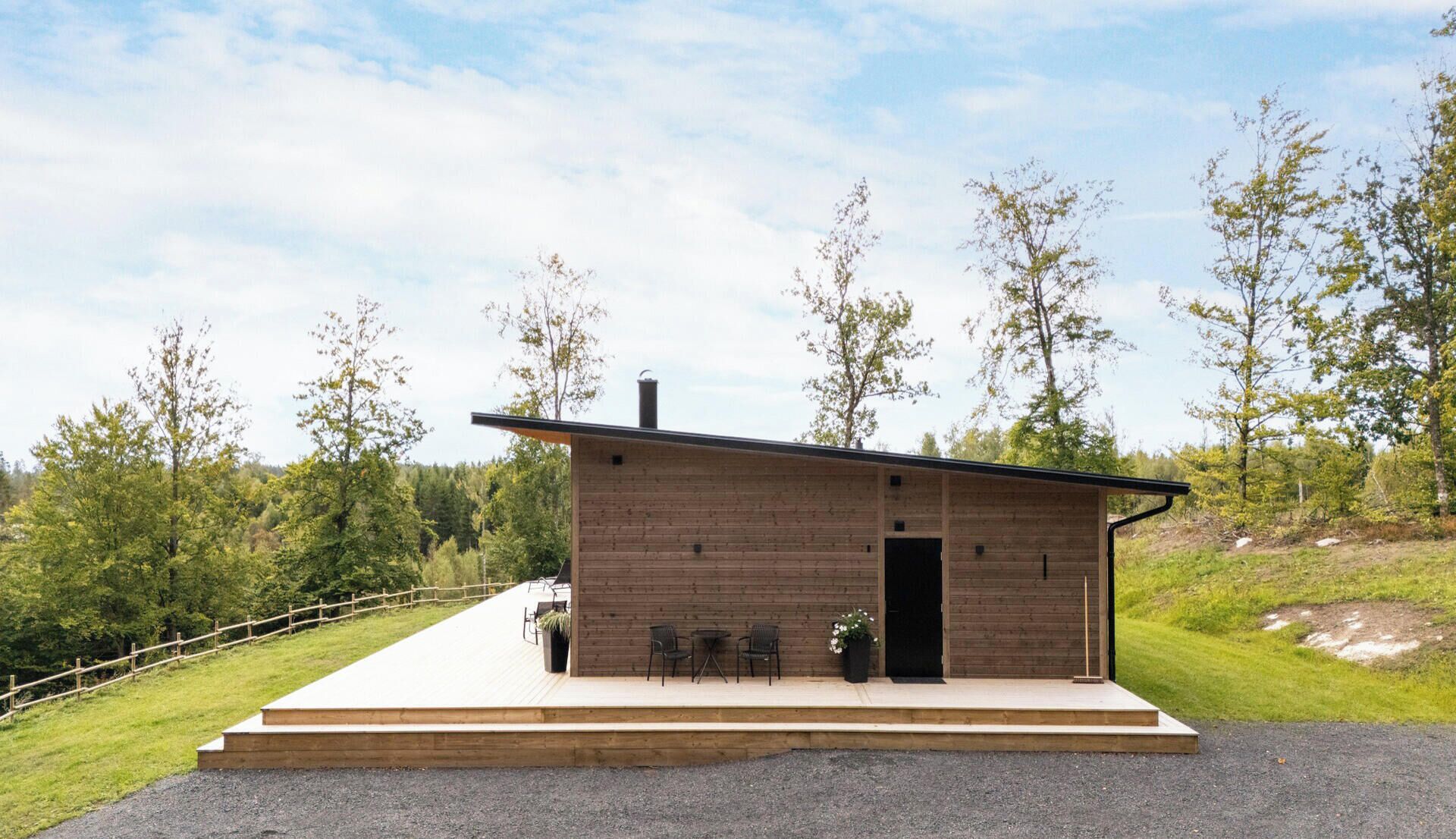 Une cabane moderne en bois, dotée d’un toit plat, d’une porte et d’un petit espace porche avec deux chaises.