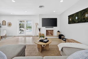 A modern living room with a fireplace, a flat-screen TV mounted on a brick wall, and a large window overlooking a pool.