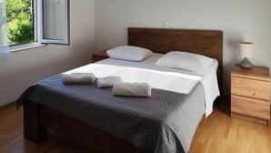 A neatly made bed with a grey bedspread and white linens, a wooden headboard, a bedside table with a lamp, and a window with a view of greenery.