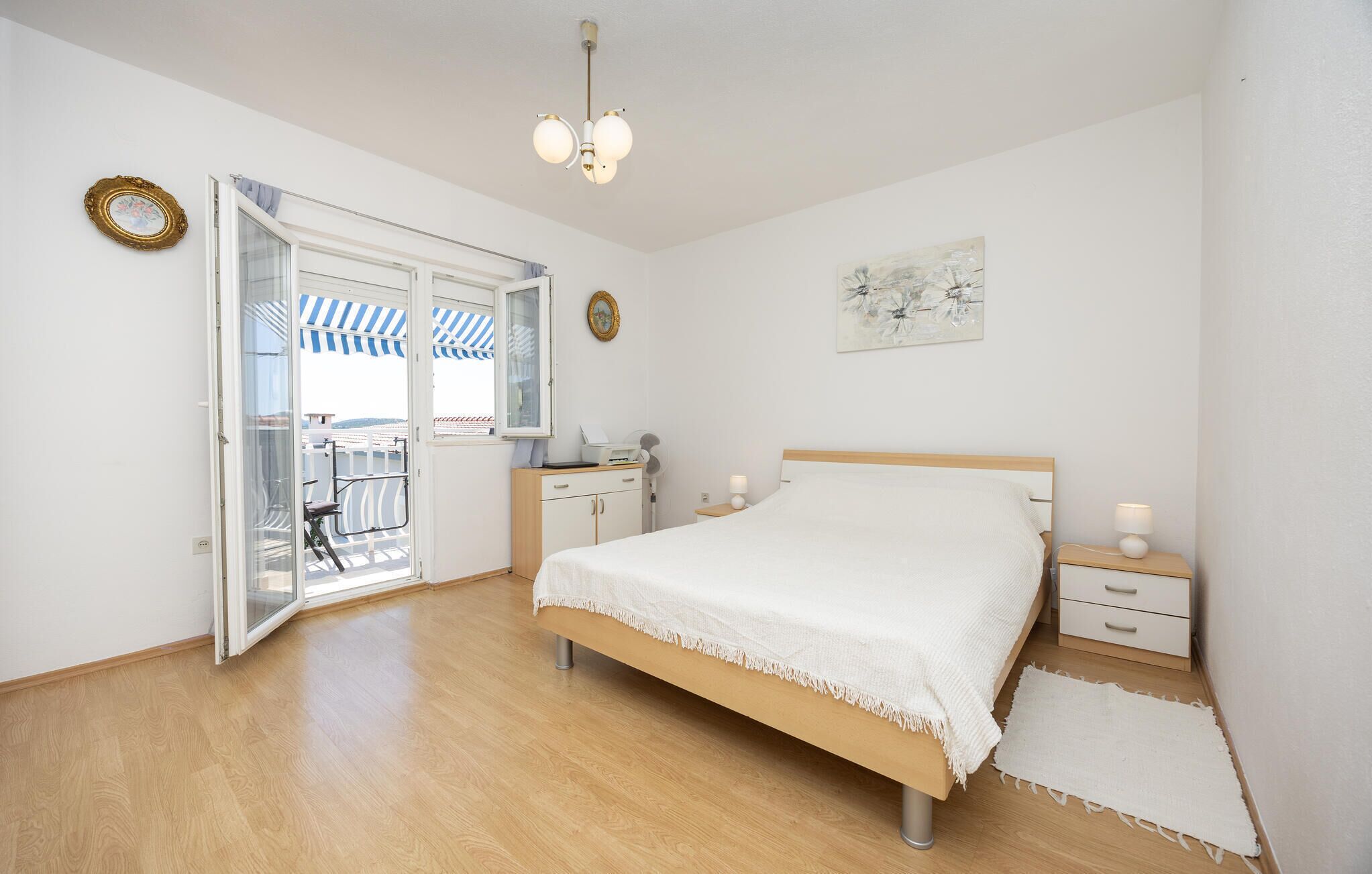 Ein Schlafzimmer mit Bett, Nachttischen, Schreibtisch, Stuhl, Kleiderschrank und einer Tür, die auf einen Balkon mit Streifenveranda führt.
