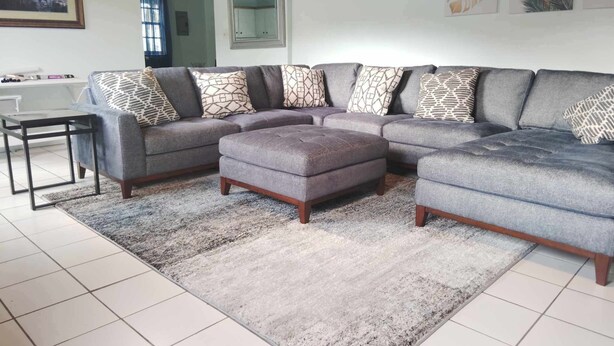 A modern living room with a grey sectional sofa, patterned throw pillows, a grey ottoman, and a large area rug. - Experience a spacious three-bedroom, two-bathroom villa on the lush Northside. (St Thomas)