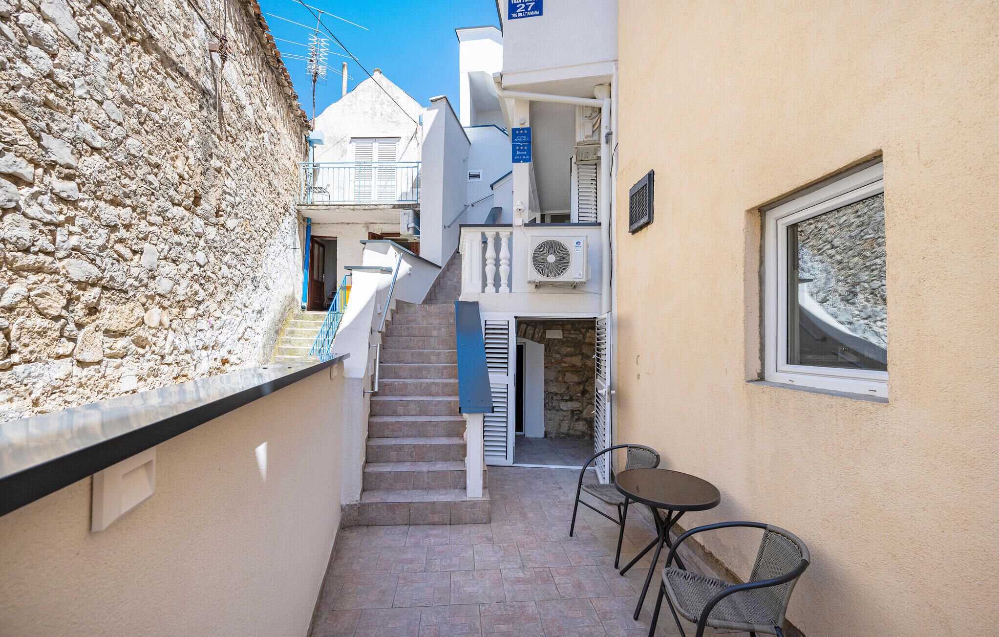 Une ruelle étroite, bordée d’un côté par un mur de pierre et de l’autre par un immeuble avec un escalier menant à un balcon. Devant l’immeuble, on trouve une petite table et deux chaises.
