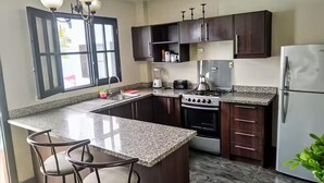 A modern kitchen with granite countertops, dark wooden cabinets, a gas stove, a refrigerator, and a window with open shutters. - La Villa Playera. (Puerto Cayo)