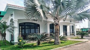 A white building with a tiled roof, large windows, and a well-maintained garden with palm trees and shrubs.