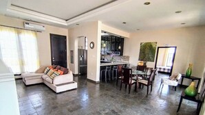 A spacious living area with a sofa, dining table, and chairs. A kitchen with appliances is visible in the background.