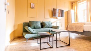 A room with a green sofa, a small table, a bed, a TV on the wall, and a picture frame.