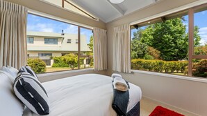 A hotel room with a large bed, beige walls, and large windows offering a view of a building and greenery outside. - Angela Close to the Lake (Taupo)