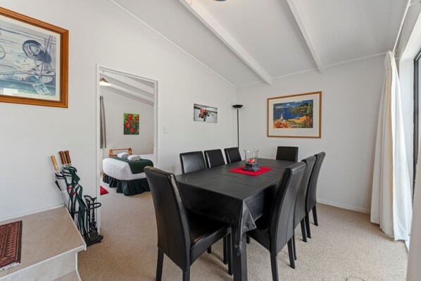 Un comedor con una mesa y sillas negras, obras de arte en las paredes y vistas a un dormitorio con cama y una silla verde.