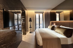 A modern hotel room with a large bed, a flat-screen TV, a desk, and a balcony with curtains.