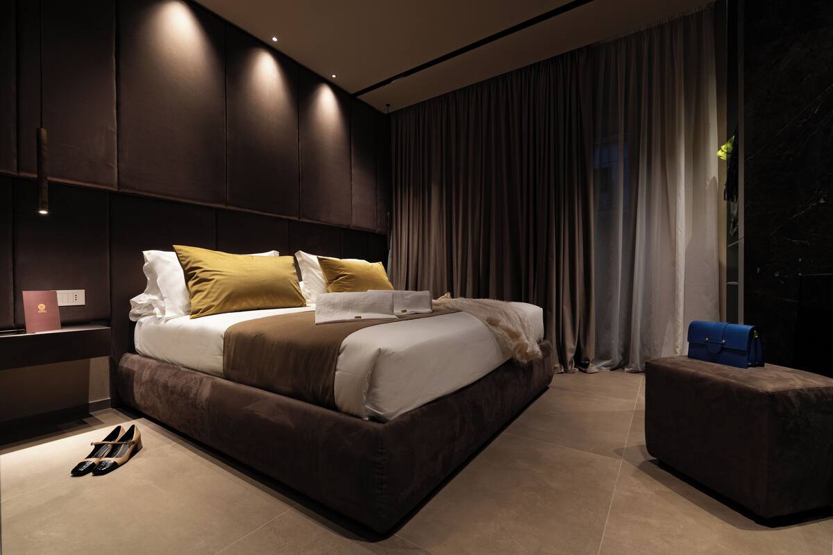 A modern bedroom with a large bed, dark wood paneling, a nightstand, and a blue bag on a small ottoman.