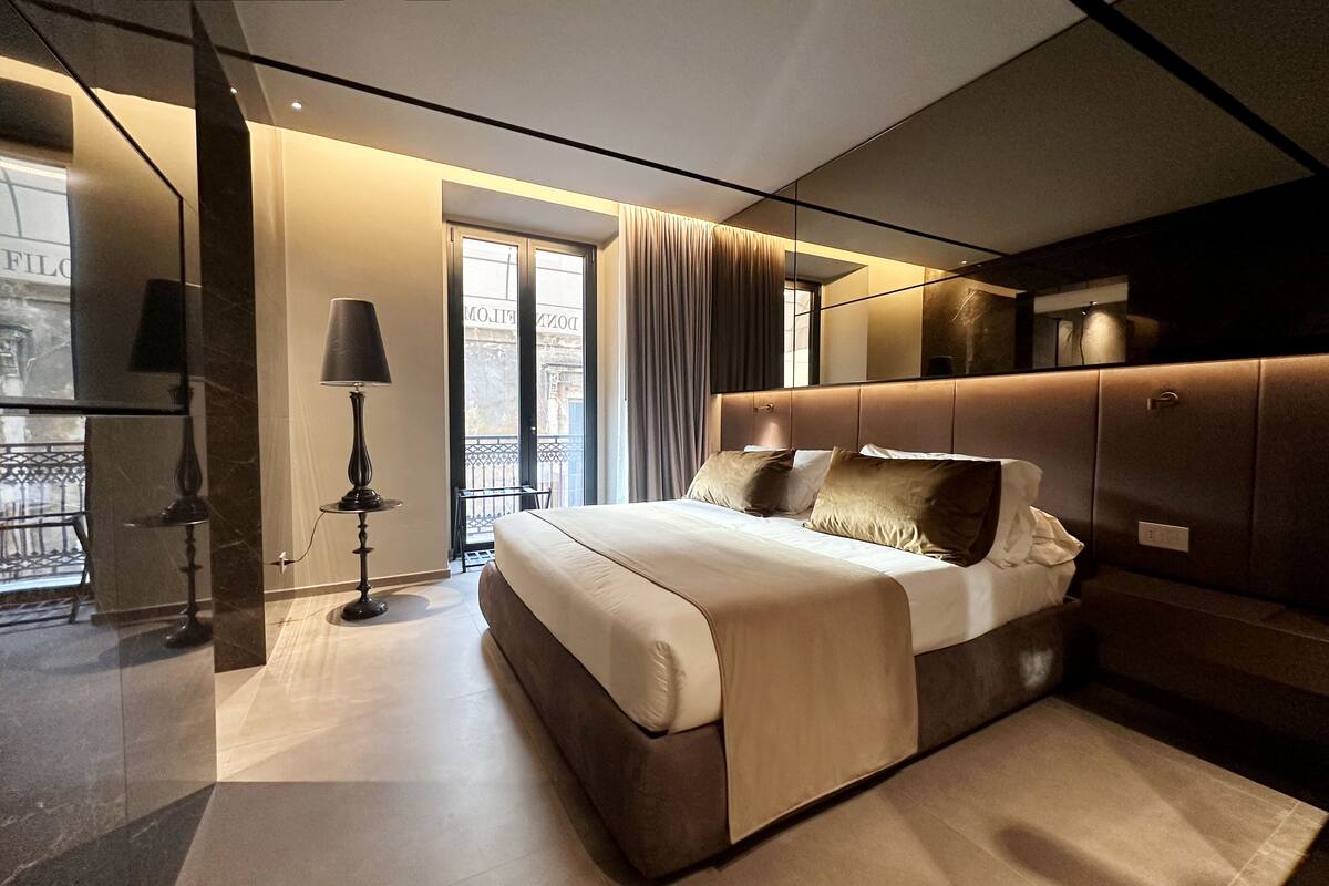 A modern bedroom with a large bed, a bedside table, a lamp, and a view of the city through a window.