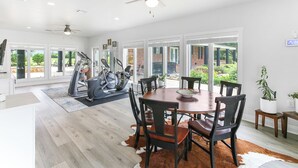 Een ruime eethoek met een grote houten tafel en stoelen, een plafondventilator en grote ramen met uitzicht op de omgeving.